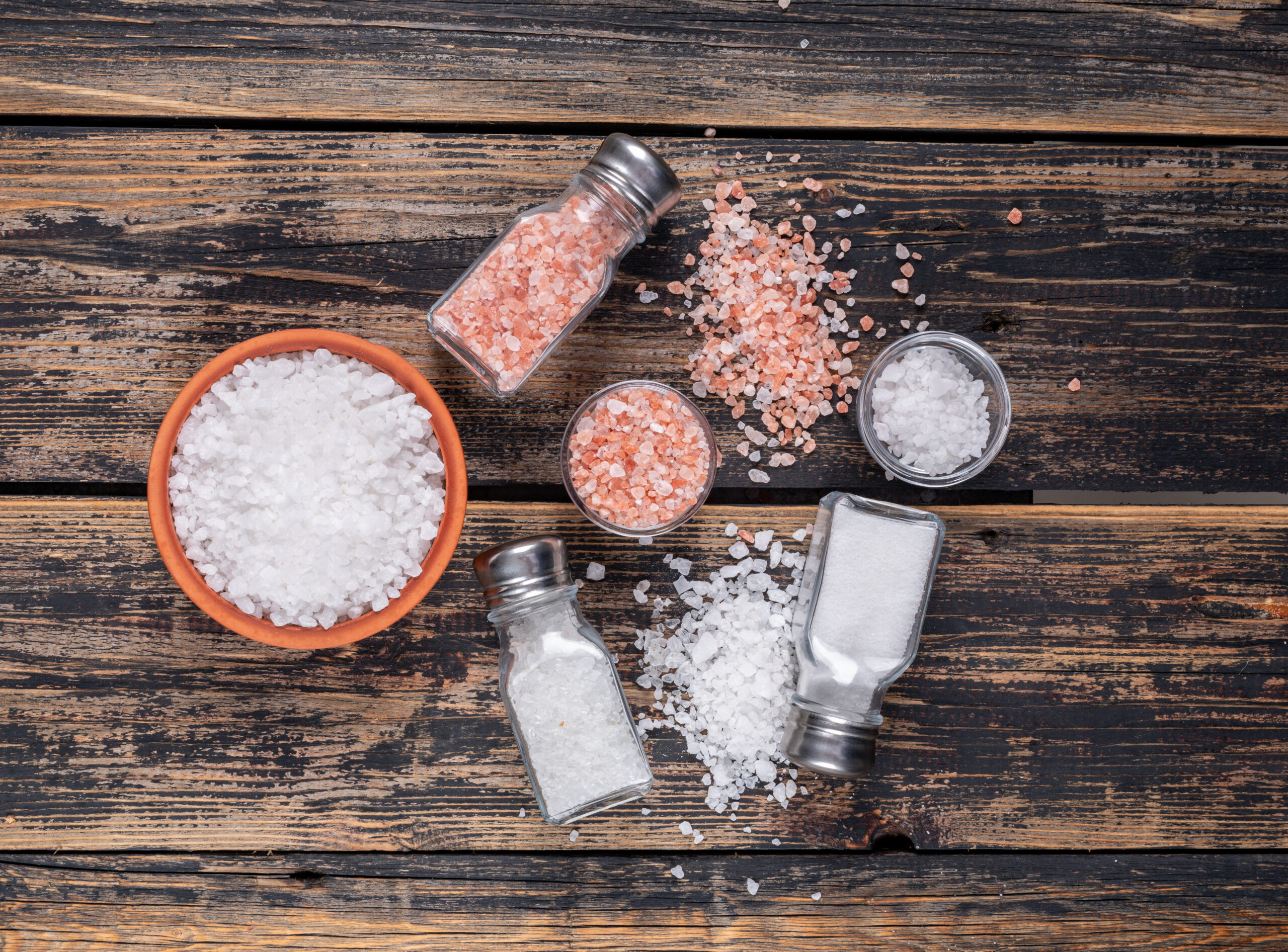 Some sea salt and himalayan salt in bowls and coming out of salt shakers on dark wooden background, flat lay.